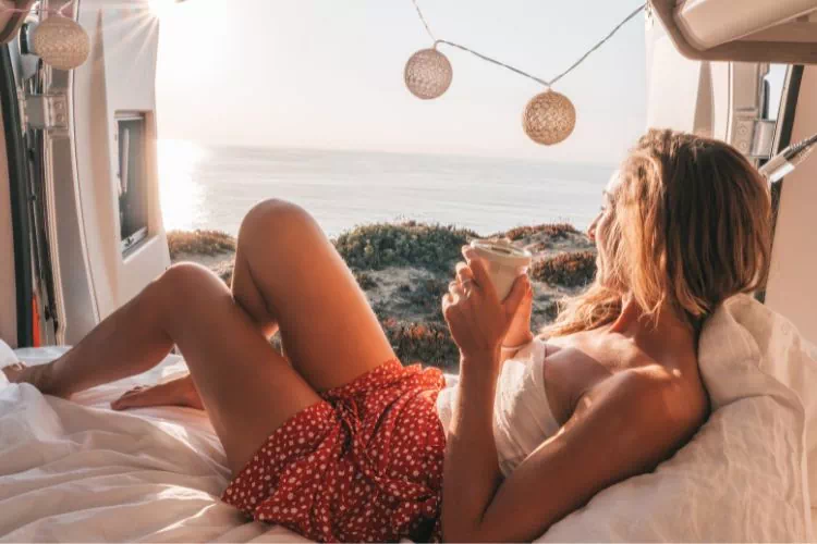 Jeune femme sur un couchage à l'arrière d'un fourgon aménagé avec vue sur la mer