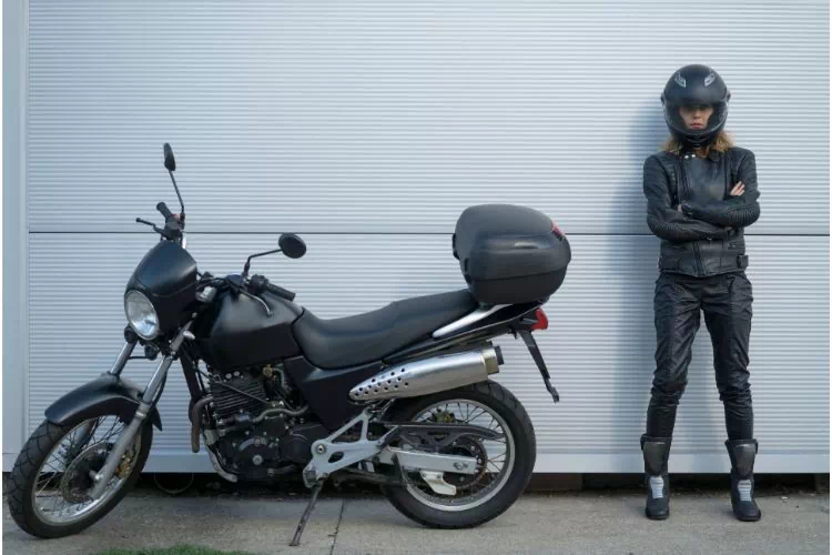Femme avec casque, blouson, pantalon et chaussures de moto à côté de sa moto